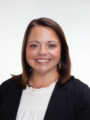 Aja Bridgewater smiling in a dark blazer and white blouse against a light gray background.