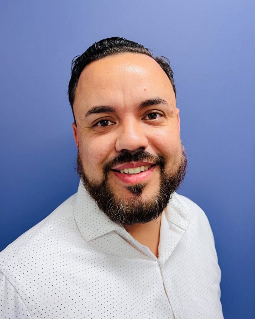 Alex Rivera smiling in a white dotted collared shirt against a blue background.