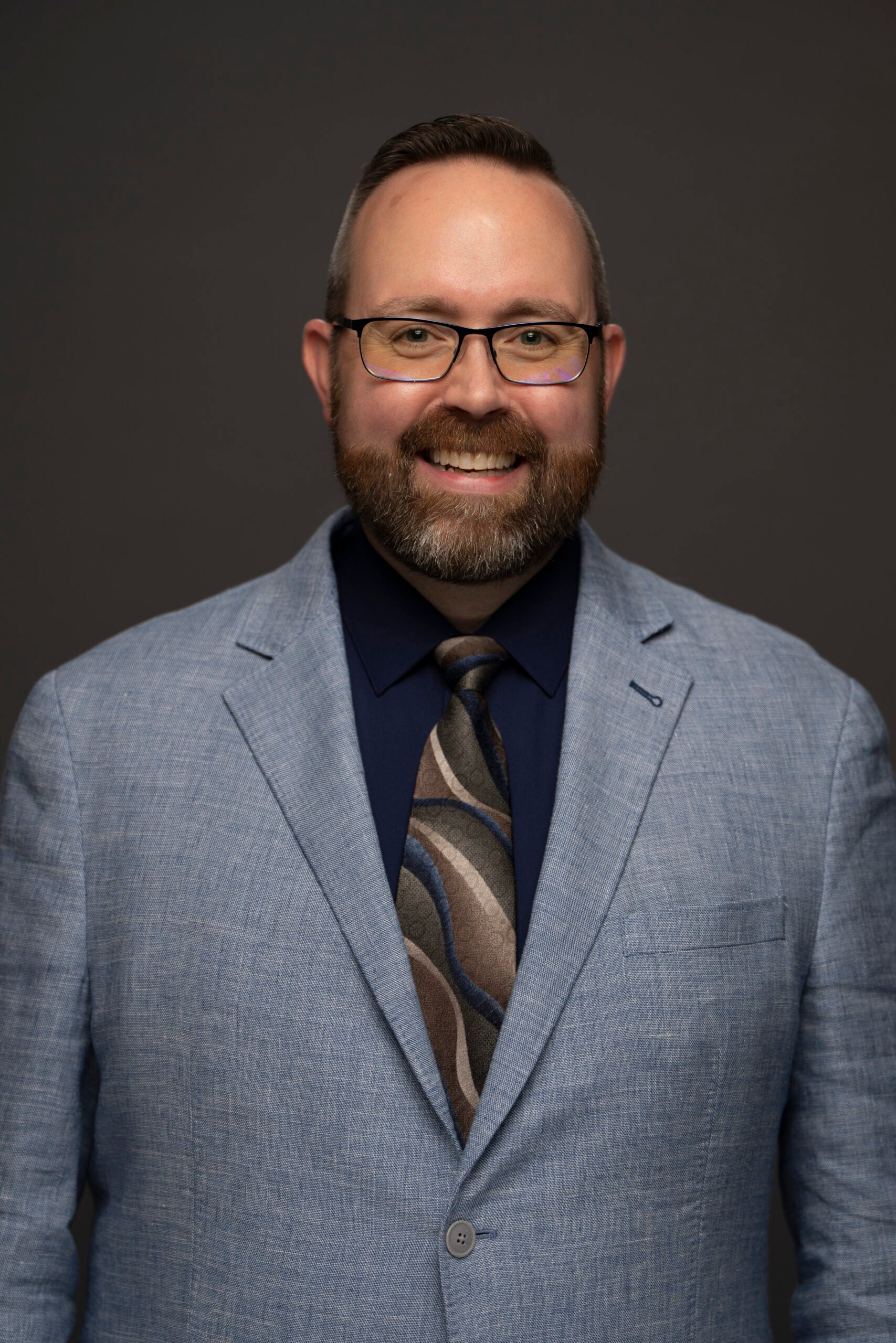 Dan Mansfield wearing glasses, a light blue suit jacket, dark shirt, and patterned tie against a dark background.