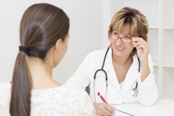 Doctor in a white coat and glasses speaking with a patient across a desk.