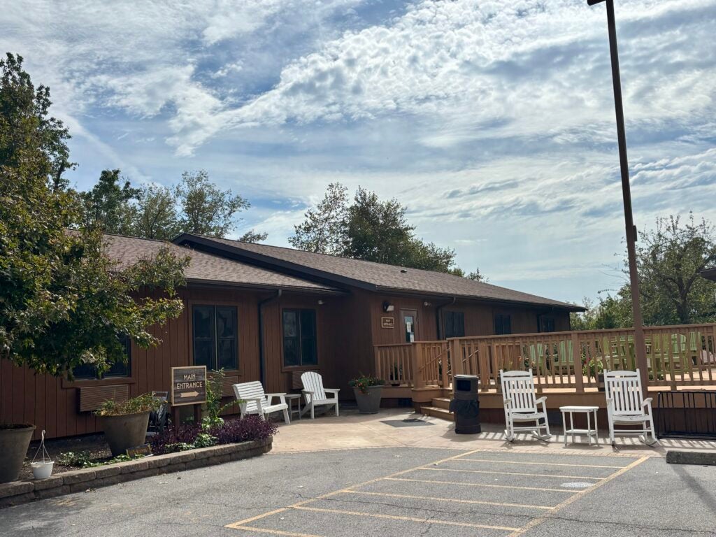 Brown single-story building with a main entrance sign, white rocking chairs, and a ramped wooden porch.