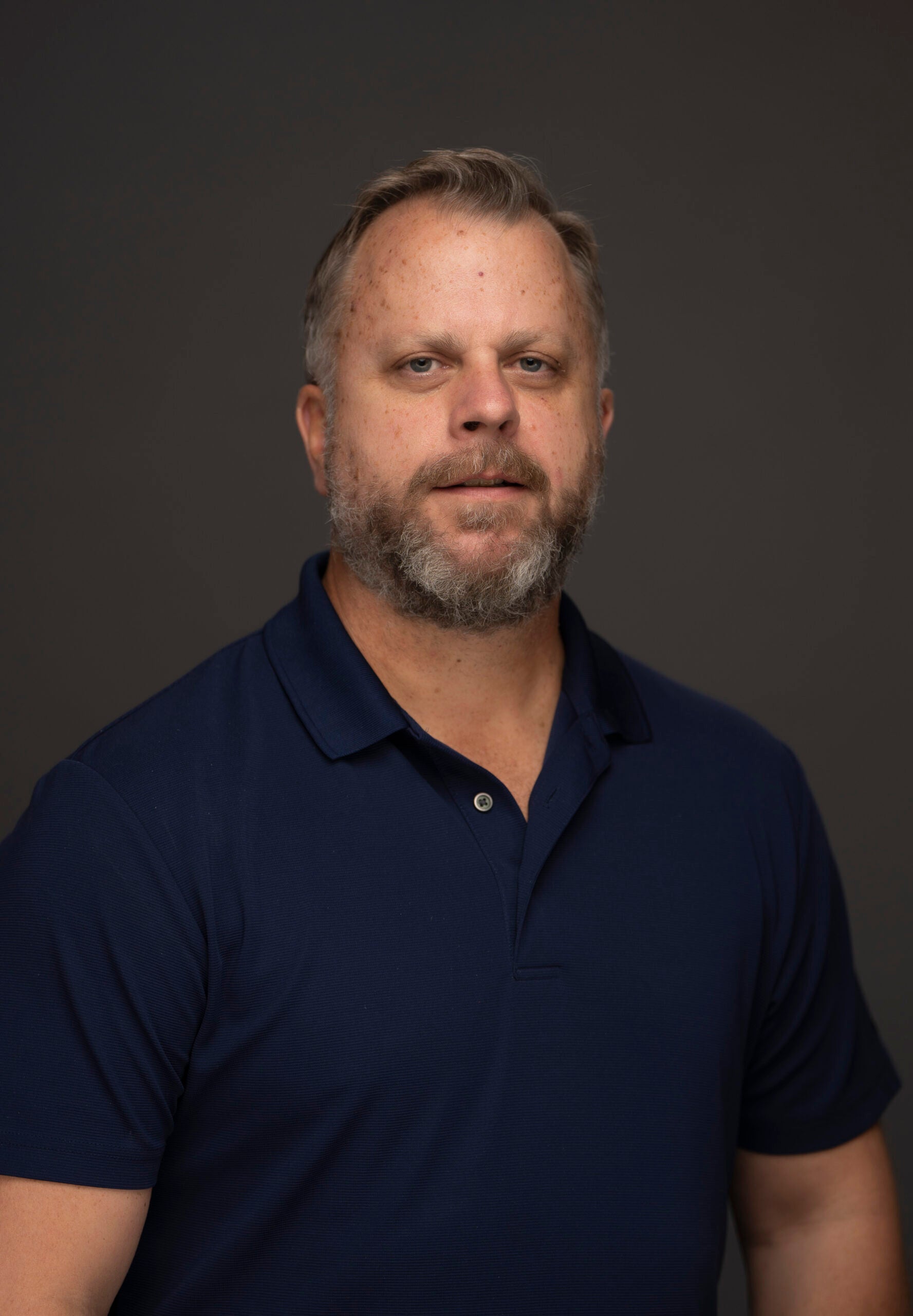 Portrait of John Sanders wearing a navy polo shirt against a dark gray background.