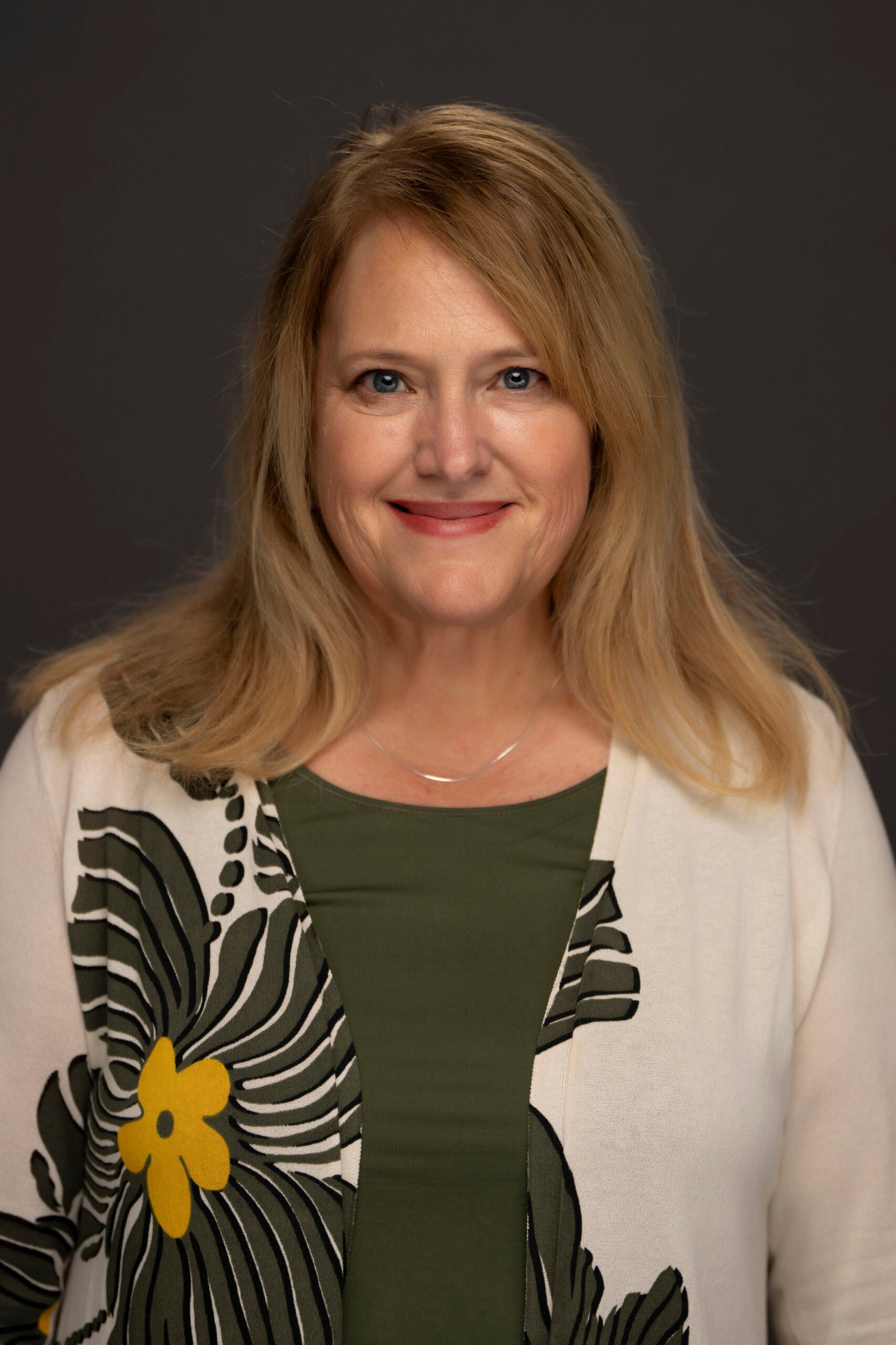 Portrait of Melissa Helm with shoulder-length blonde hair, wearing a patterned cardigan over a dark green top against a dark background.
