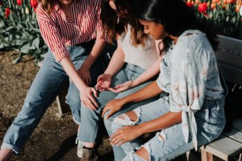 three women wearing blue denim sitting on bench outside, hands on each other in support