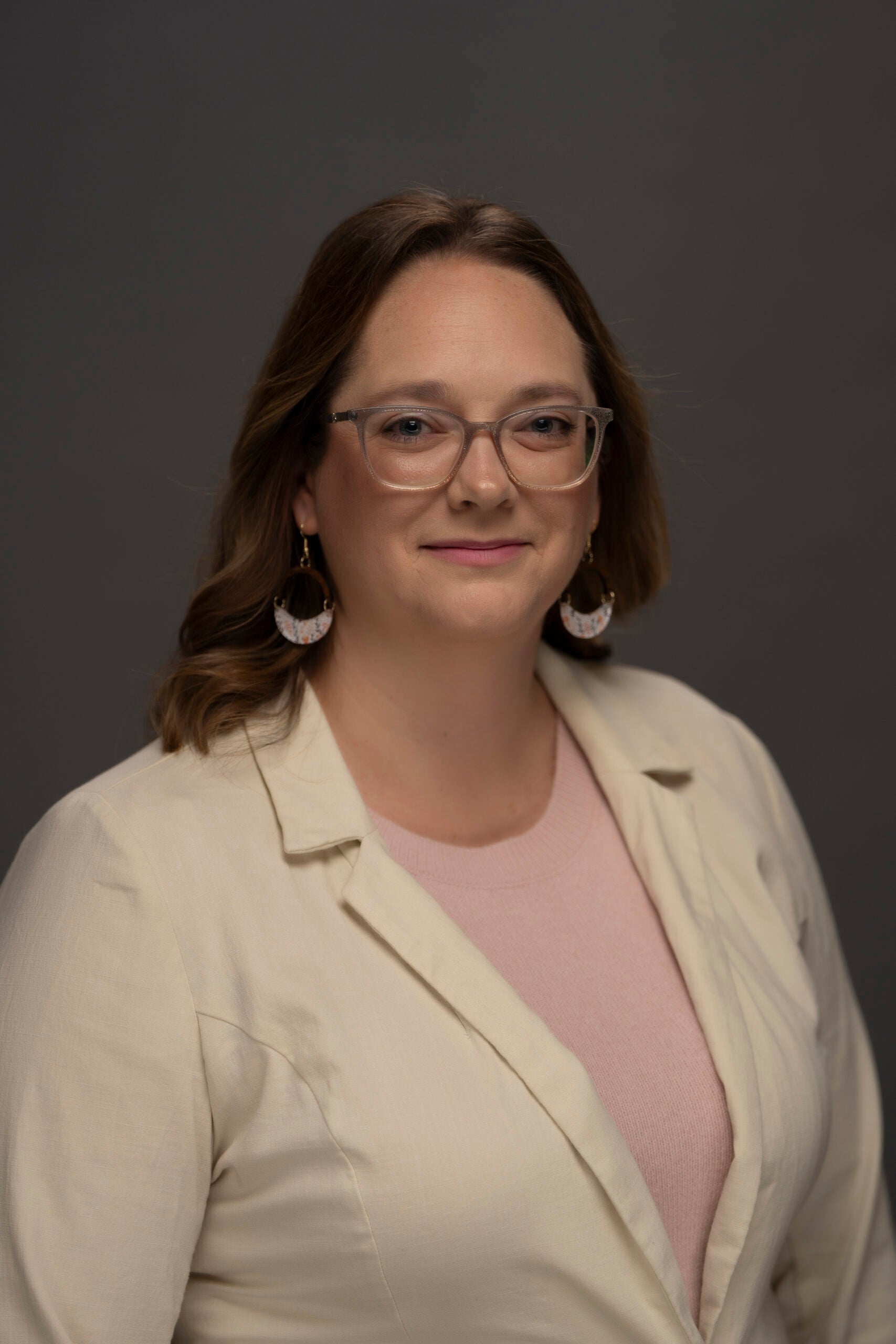 Valerie Klein wearing glasses, a cream blazer, and large earrings against a dark gray background.