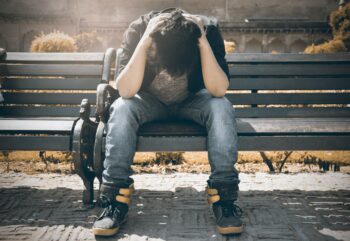 young man sitting on bench outside with head down and elbows on knees