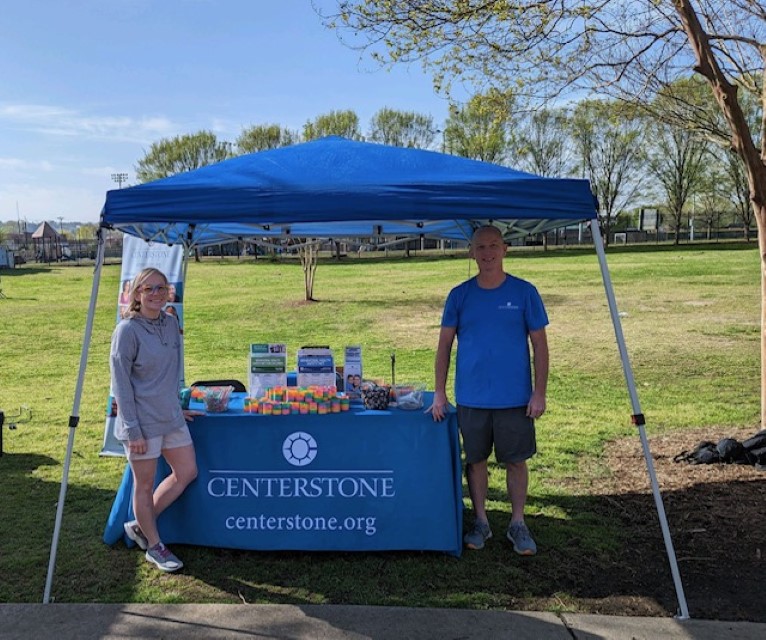Two Centerstone staff on either side of an outdoor event booth beneath a tent on a sunny day.