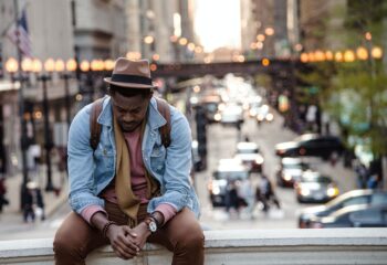 sad man wearing fedora and denim jacket looking down at folded hands outside in city