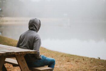 man wearing hoodie and jeans sitting on picnic bench facing out toward body of water