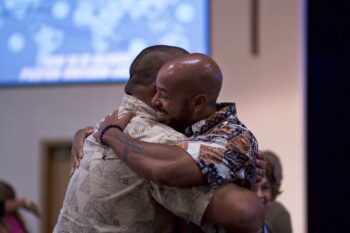 two men hugging inside, man in front smiling