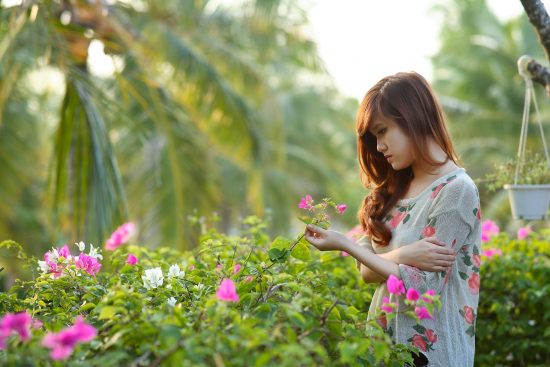 woman holding flower next to flower bushes with palm trees in background