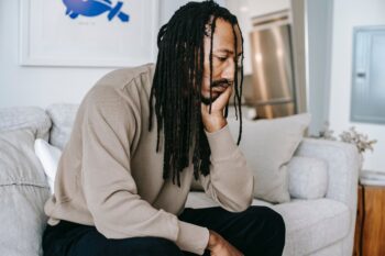 sad man with dreadlocks and septum ring sitting on sofa, chin resting on hand