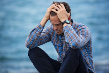 man wearing watch sitting in front of water with hands on head distressed